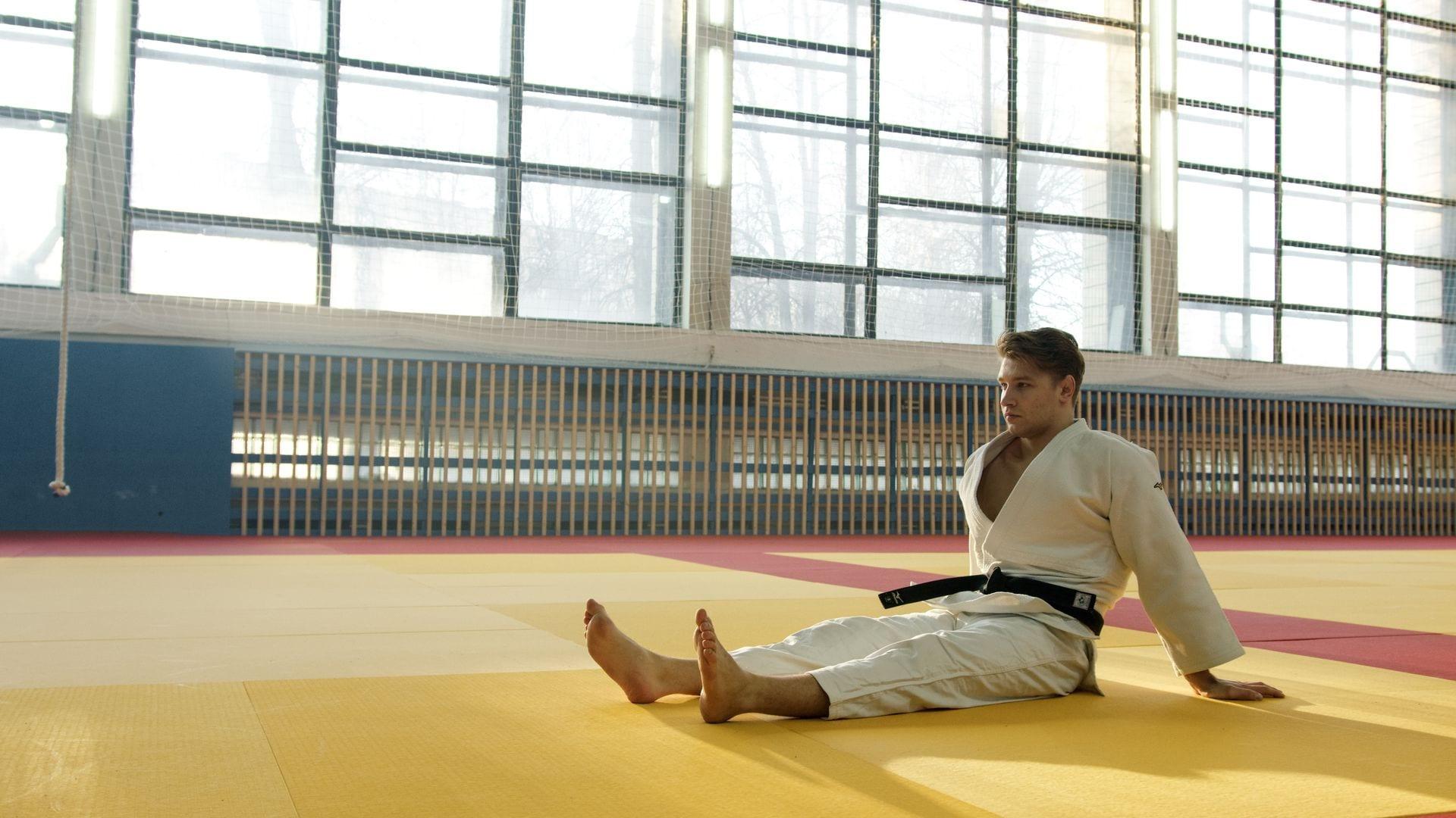 A judoka in a white gi and black belt sitting on the mat in a large, sunlit training hall